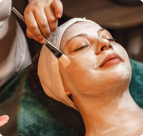 A woman lying comfortably while a technician applies facial treatment with a brush, promoting skin care and relaxation